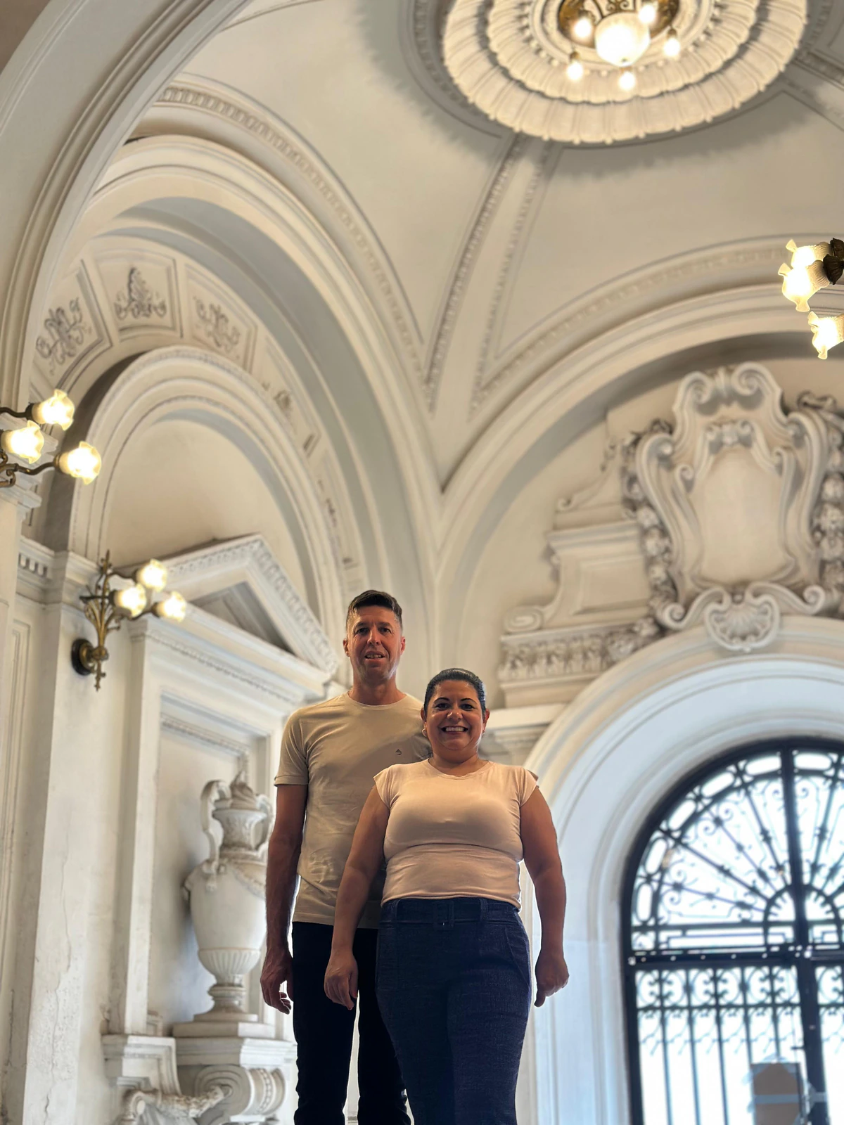 Alline e Anderson sorrindo lado a lado num salão de arcos brancos com ornamentos em relevo e lustres acesos