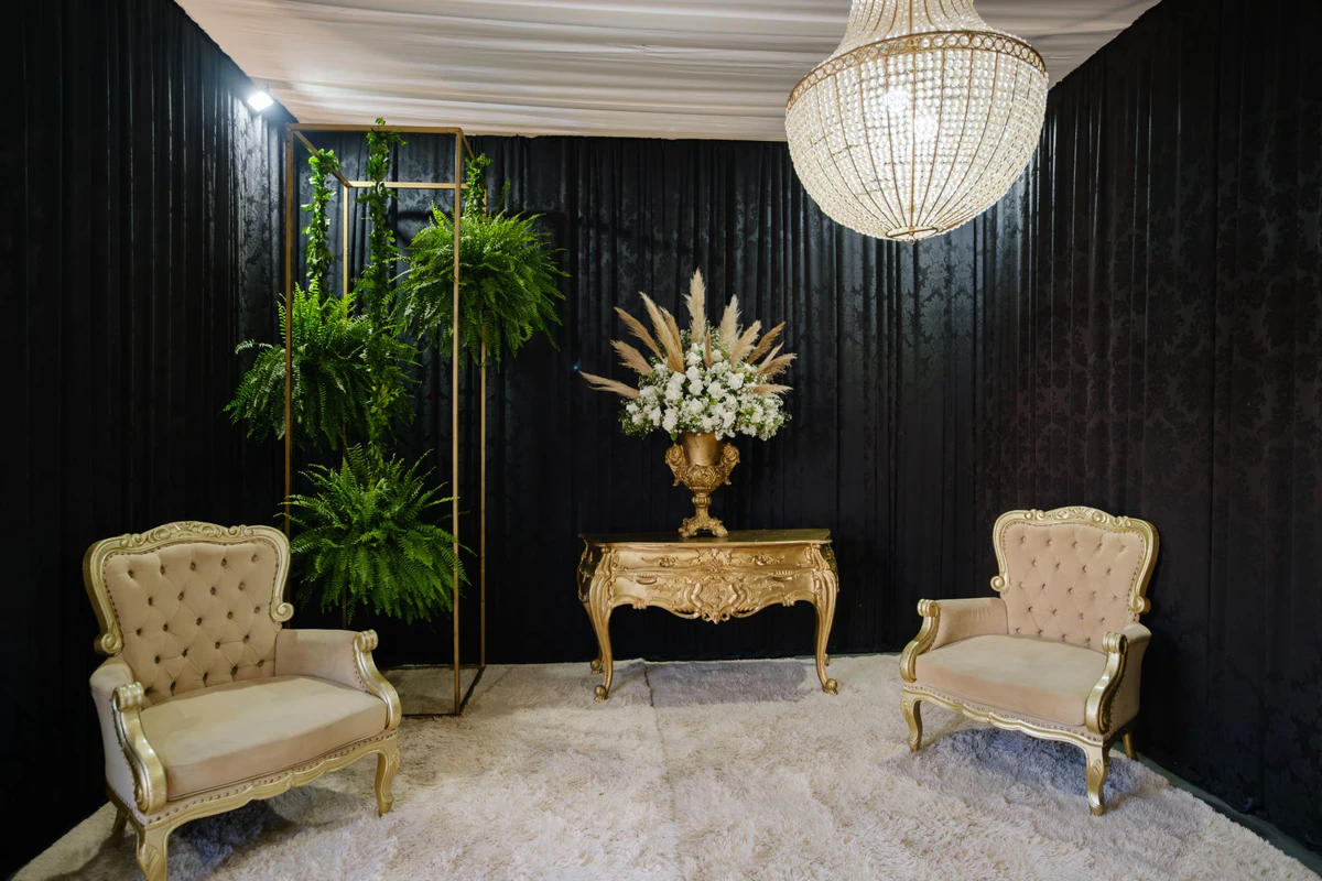Cantinho de casamento com cortina preta, lustre de cristal, aparador dourado entalhado, ânfora de flores brancas e poltronas capitonê
